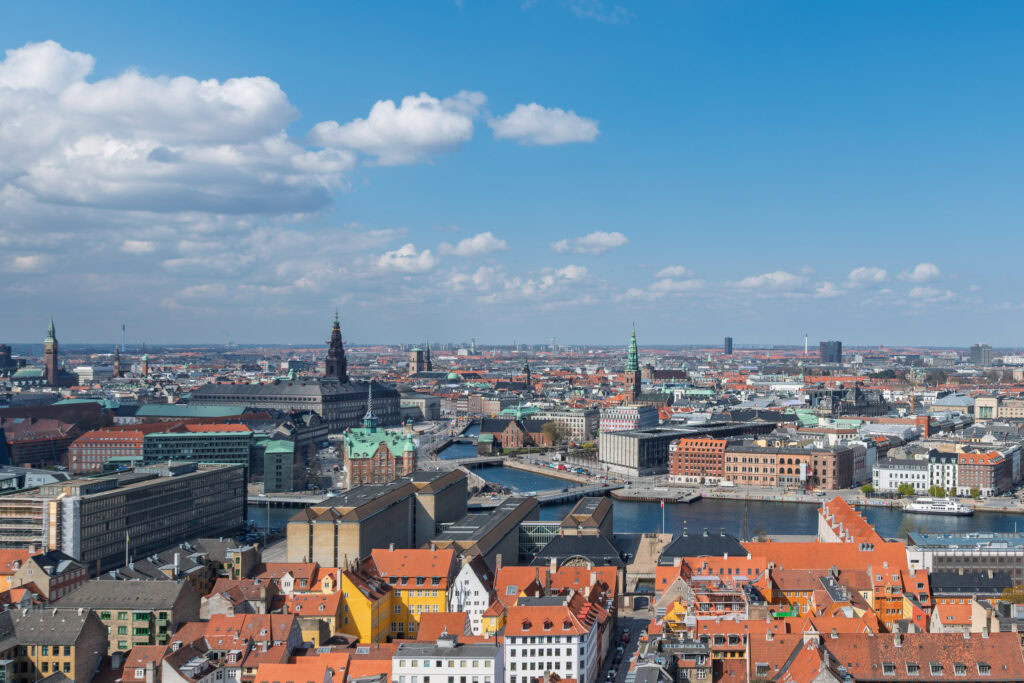 Aerial view of central Copenhagen on a partly cloudy day, showing the Christianshavn canal, terracotta rooftops, copper-patinated spires, and the city skyline stretching to the horizon under a blue sky.