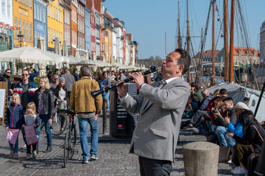 A man playing a clarinet on a busy harbour promenade with colourful buildings and boats.