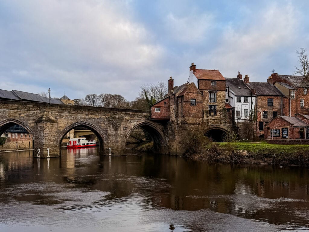 A stone bridge over a river with historic buildings along the bank under a cloudy sky.