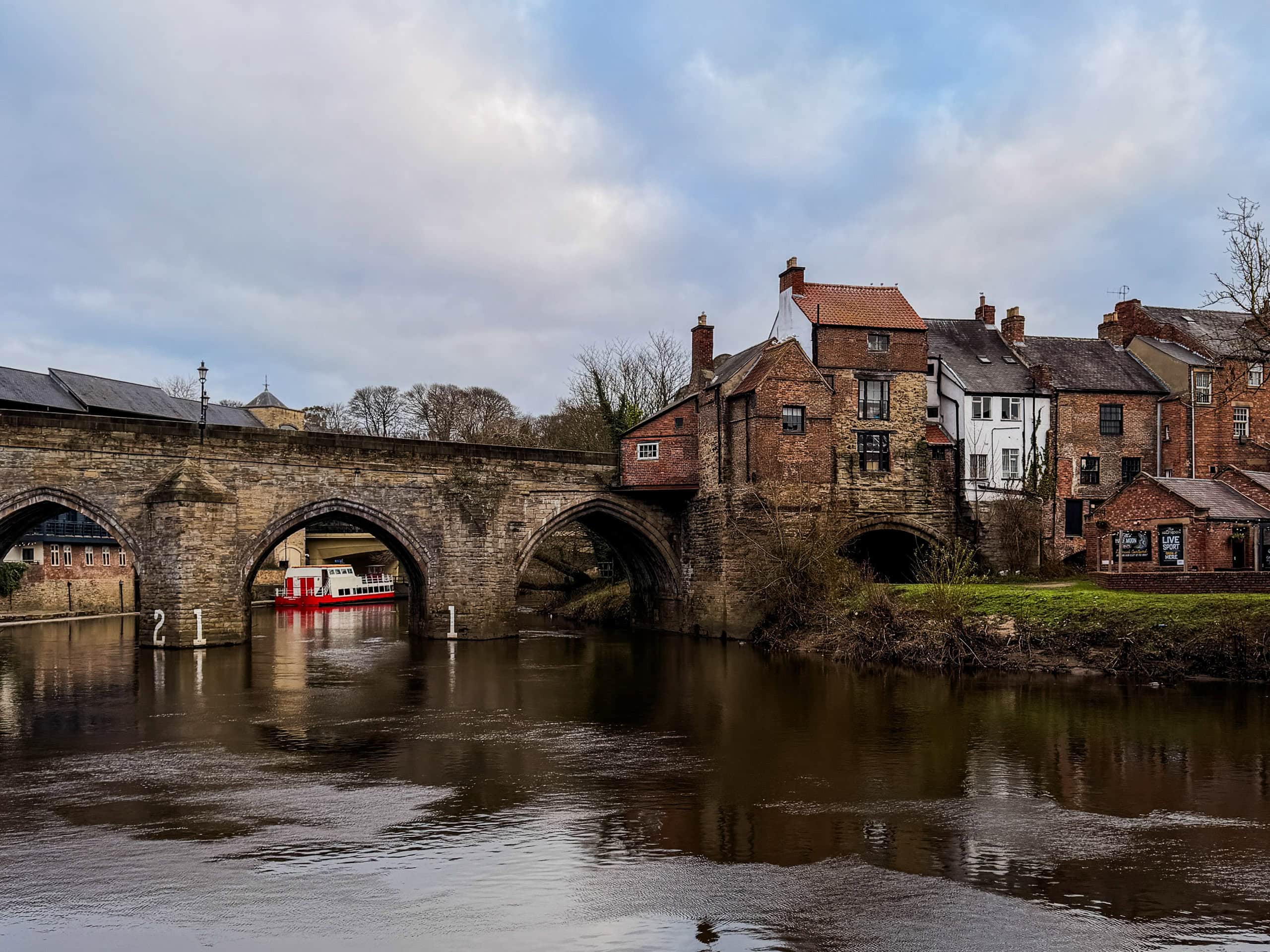 A stone bridge over a river with historic buildings along the bank under a cloudy sky.