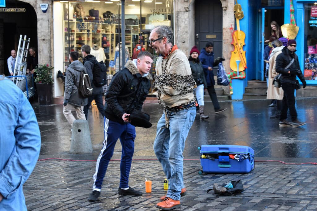 Street performer wrapped in heavy chains entertaining a crowd on a busy Edinburgh street, while a volunteer interacts during the act. Wet cobbled pavement, colourful shopfronts, and passing pedestrians create a lively urban atmosphere.