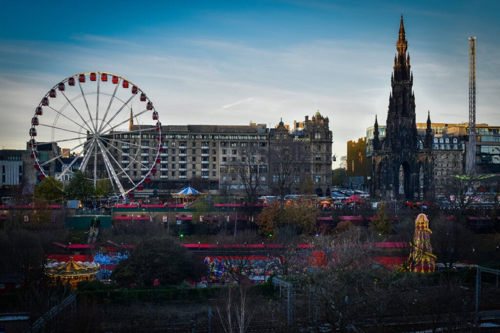 Edinburgh skyline at dusk featuring the Scott Monument beside a large Ferris wheel and festive market stalls below. Historic city architecture contrasts with winter fairground lights under a calm blue evening sky.