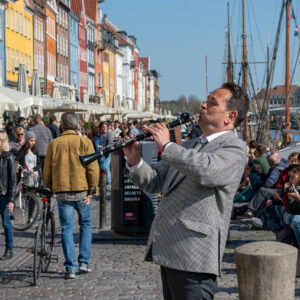 A man playing a flute on a busy harbour promenade with colourful buildings and boats in the backgrou.