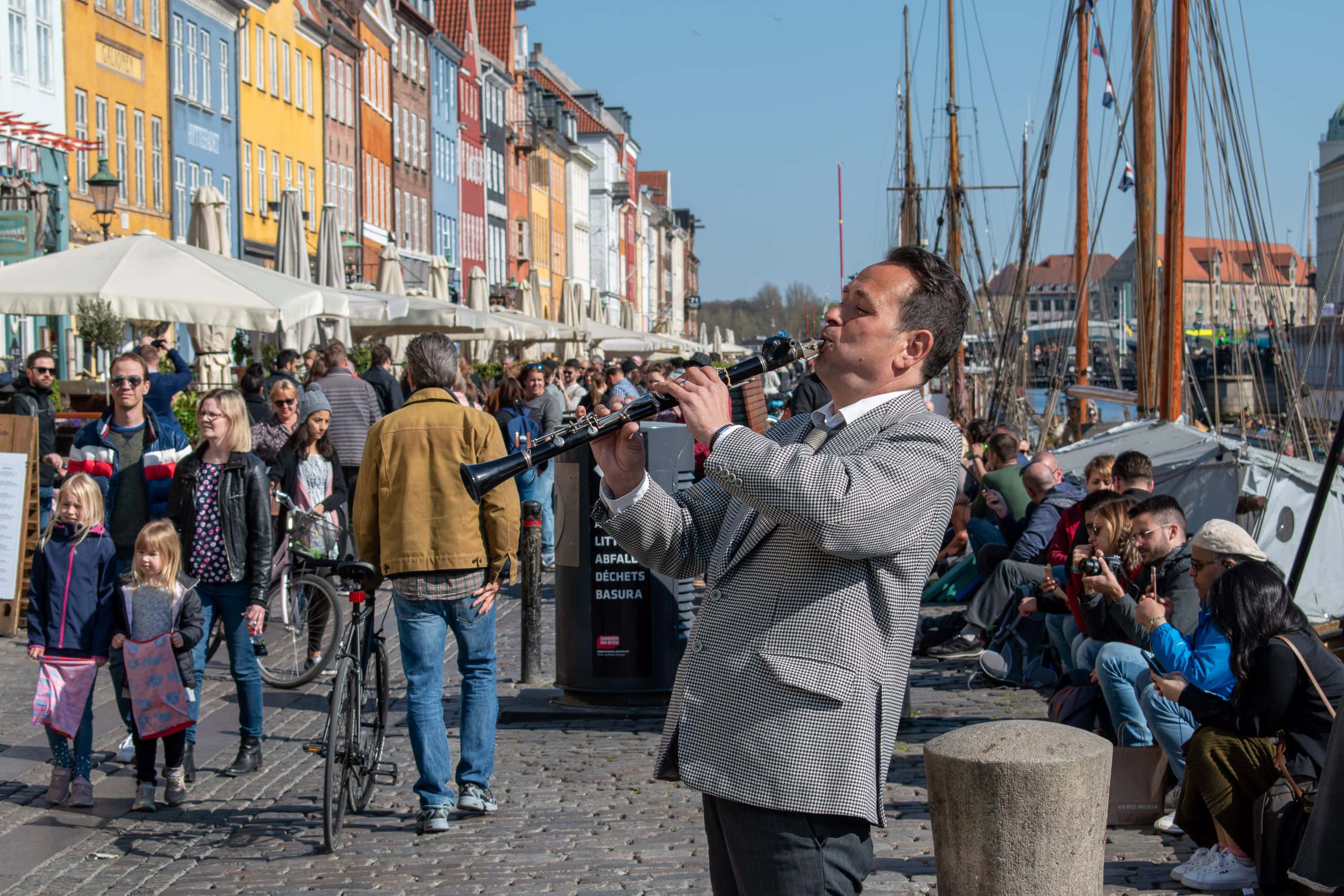 A man playing a flute on a busy harbour promenade with colourful buildings and boats in the backgrou.
