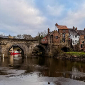 A quaint riverside scene with historic stone bridge and old brick houses under cloudy sky.